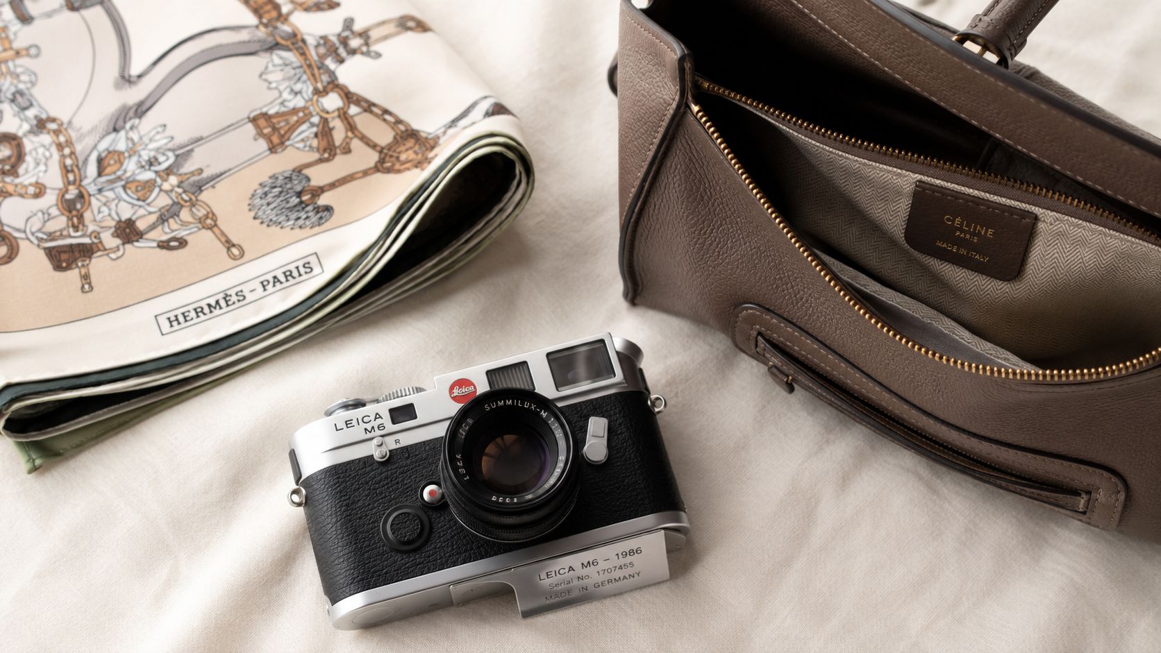 A vintage Leica M6 camera sits beside a designer handbag and an Hermes scarf on white fabric.
