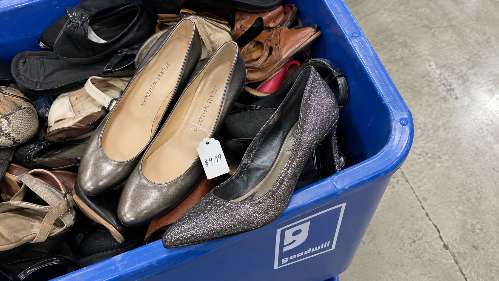 A bin filled with assorted used women's shoes at a Goodwill store, including prominent dark silver heels.